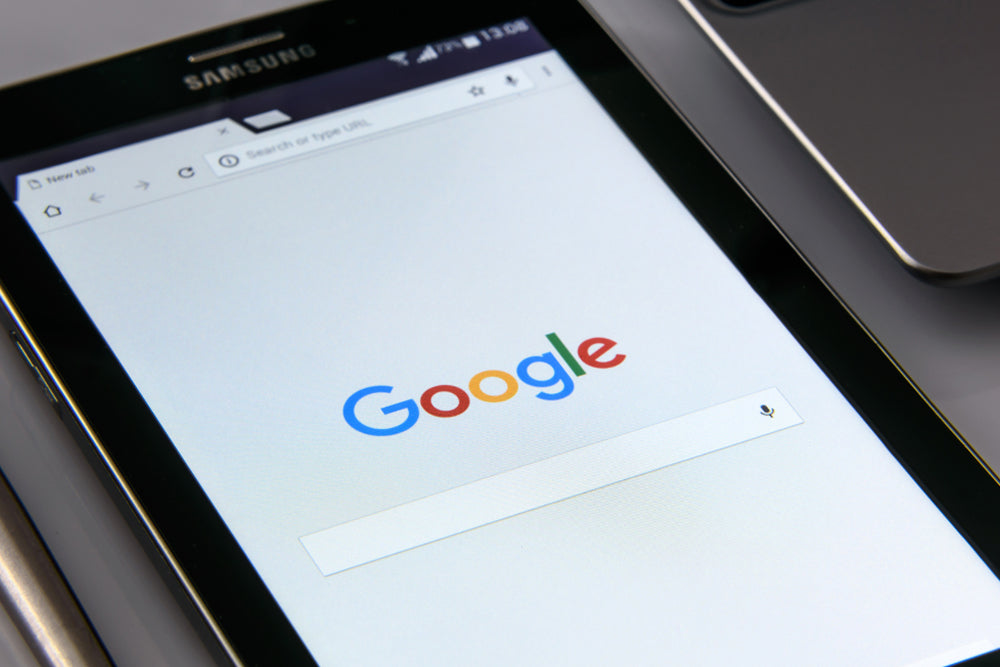 A close-up of a black Samsung tablet displaying the Google search homepage on its screen, with part of a laptop visible in the background.