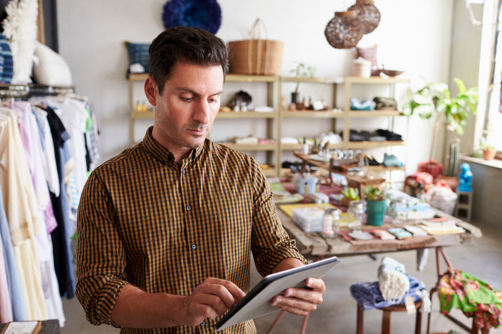 A man in a brown and yellow checkered shirt stands in a retail store holding a tablet, with clothing racks and shelves visible in the background.