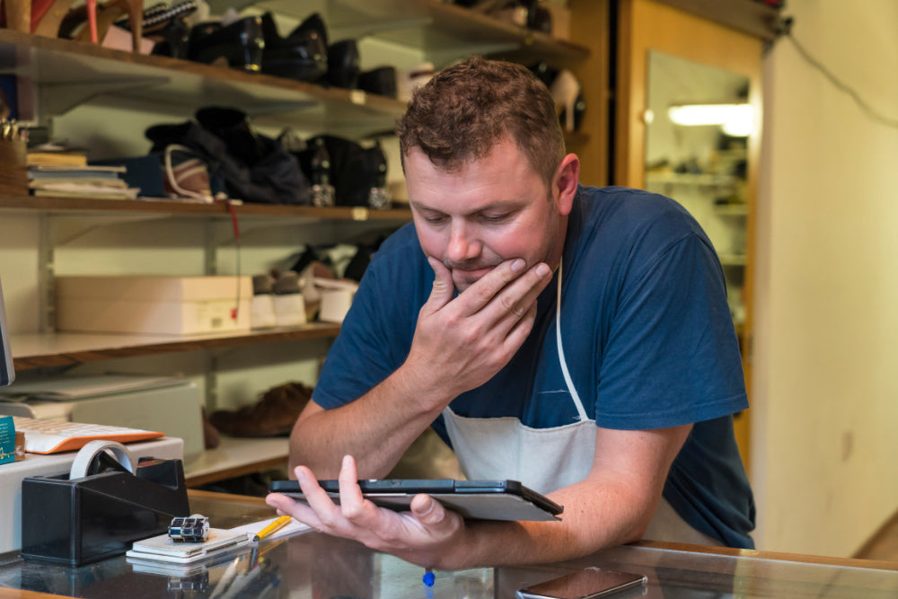 A man wearing a blue t-shirt and white apron looks down at a tablet on a counter inside a shoe store with shelves of shoes in the background.