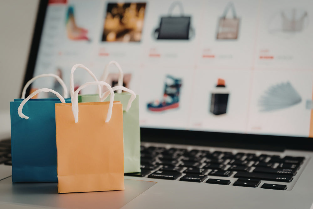 Three colorful paper shopping bags in blue, orange, and light green sit on a laptop keyboard, with a blurred online shopping storefront visible on the screen behind them.
