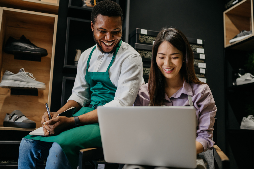 A smiling store employee in a green apron writes in a notebook while a woman looks at a laptop in a shoe store.